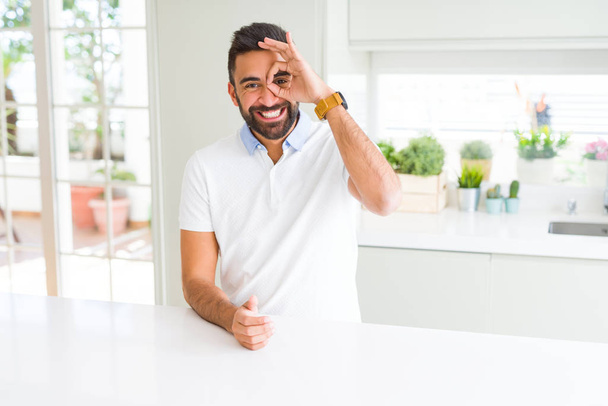 schöner hispanischer Mann legeres weißes T-Shirt zu Hause tut ok Geste mit lächelnden Händen, Augen durch Finger mit glücklichem Gesicht. - Foto, Bild