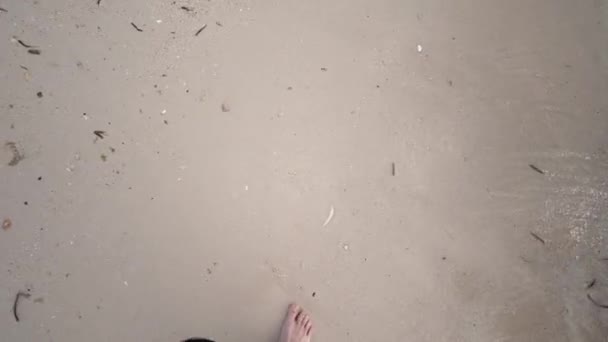 Een close-up eerste persoon perspectief van een jonge Kaukasische mannen benen als hij loopt op een zandstrand met golven af en toe wassen boven zijn blote voeten krijgen ze nat en bedekt ze met zand. - Video