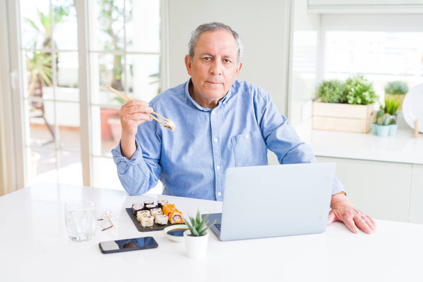 Komea liike vanhempi mies syö toimitus sushi työskennellessään kannettavan kanssa luottavainen ilme älykäs kasvot ajattelu vakava
 - Valokuva, kuva