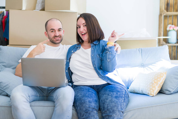 Νεαρό ζευγάρι κάθεται στον καναπέ arround κουτιά από χαρτόνι κινείται σε ένα νέο σπίτι χρησιμοποιώντας φορητό υπολογιστή πολύ χαρούμενος δείχνοντας με το χέρι και το δάχτυλο στο πλάι - Φωτογραφία, εικόνα