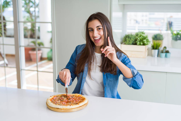 schöne junge Frau schneidet eine leckere Pizzascheibe mit einem Ausstecher überrascht mit einer Idee oder Frage Zeigefinger mit glücklichem Gesicht, Nummer eins - Foto, Bild
