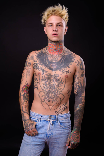 Студійний знімок молодого красивого бунтівного чоловіка з світлим волоссям та татуюваннями на тілі без сорочки на чорному тлі
 - Фото, зображення