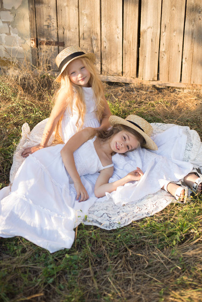 Cute little girls with blond long hair in a summer field at sunset in white dresses with a straw hat - Photo, Image