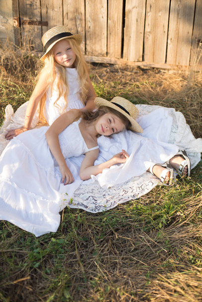 Cute little girls with blond long hair in a summer field at sunset in white dresses with a straw hat - Фото, изображение