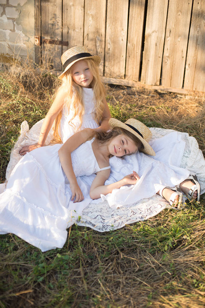 Cute little girls with blond long hair in a summer field at sunset in white dresses with a straw hat - Фото, изображение