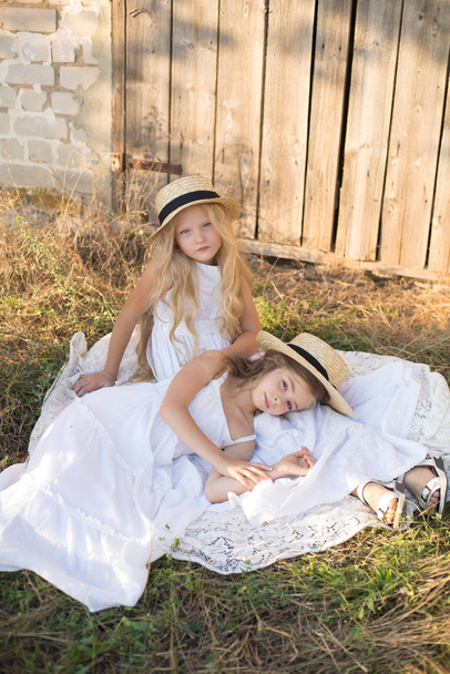 Cute little girls with blond long hair in a summer field at sunset in white dresses with a straw hat - 写真・画像