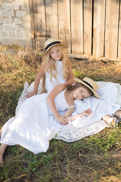 Cute little girls with blond long hair in a summer field at sunset in white dresses with a straw hat - Fotó, kép