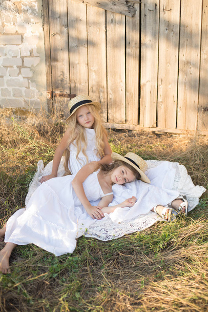 Cute little girls with blond long hair in a summer field at sunset in white dresses with a straw hat - Zdjęcie, obraz