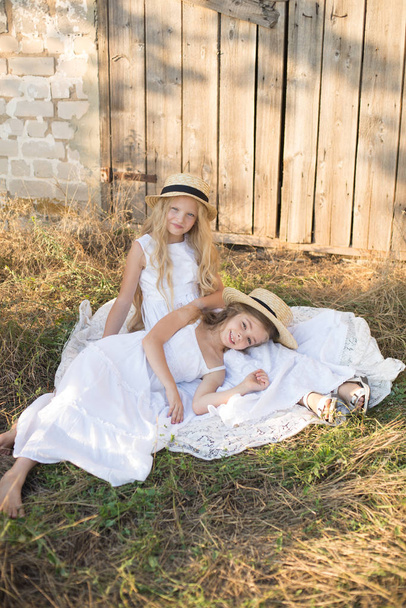 Cute little girls with blond long hair in a summer field at sunset in white dresses with a straw hat - Foto, Imagem