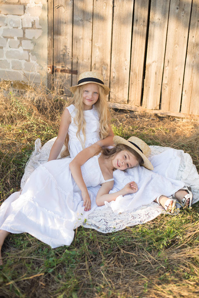 Cute little girls with blond long hair in a summer field at sunset in white dresses with a straw hat - Foto, imagen