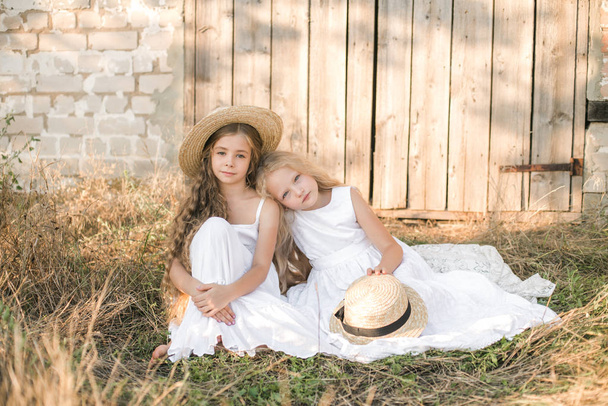 Cute little girls with blond long hair in a summer field at sunset in white dresses with a straw hat - Fotoğraf, Görsel