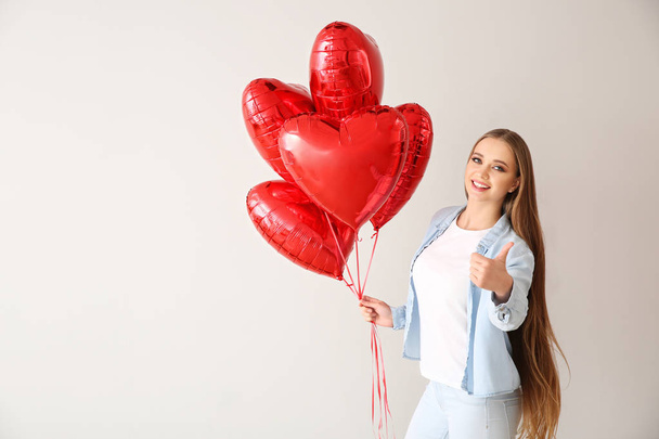 Beautiful young woman with heart-shaped air balloons showing thumb-up on light background - Foto, afbeelding
