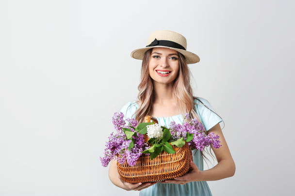 Mooie jonge vrouw met boeket van lila bloemen in mand op lichte achtergrond - Foto, afbeelding