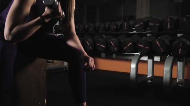 young sports girl engaged in the evening in the gym. Does exercise lifting dumbbells sitting. - Footage, Video