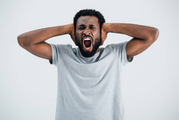 συναισθηματικός Αφρικανός Αμερικανός άντρας με γκρι μπλουζάκι ουρλιάζοντας και κλείνοντας αυτιά απομονωμένα σε γκρι - Φωτογραφία, εικόνα