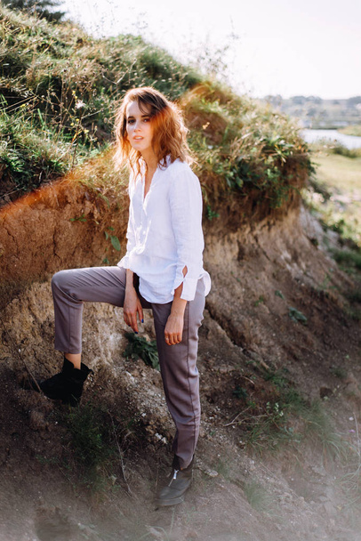 Κομψό θηλυκό μοντέλο σε πουκάμισο και παντελόνια που ποζάρει με το πόδι στο λόφο - Φωτογραφία, εικόνα