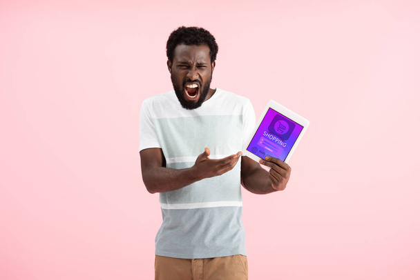 συναισθηματικός Αφρικανός Αμερικανός άνθρωπος ουρλιάζοντας και δείχνοντας ψηφιακό δισκίο με την εφαρμογή αγορών, απομονώνονται σε ροζ - Φωτογραφία, εικόνα