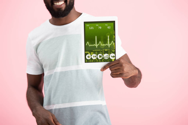bijgesneden weergave van glimlachend Afro-Amerikaanse man toont digitale tablet met gezondheid app, geïsoleerd op roze - Foto, afbeelding