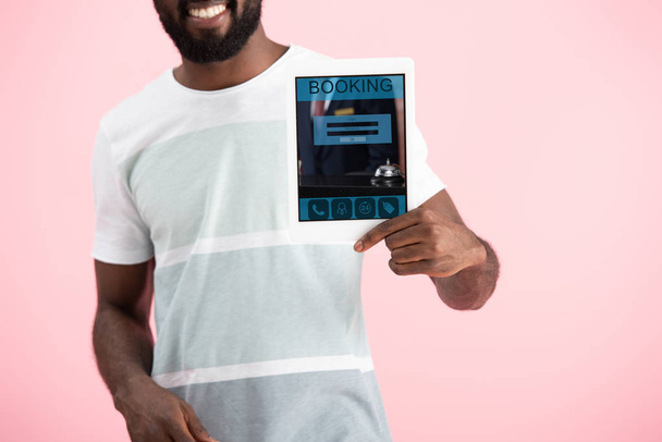 ピンクに隔離された予約アプリでデジタルタブレットを示す笑顔のアフリカ系アメリカ人男性のトリミングビュー - 写真・画像