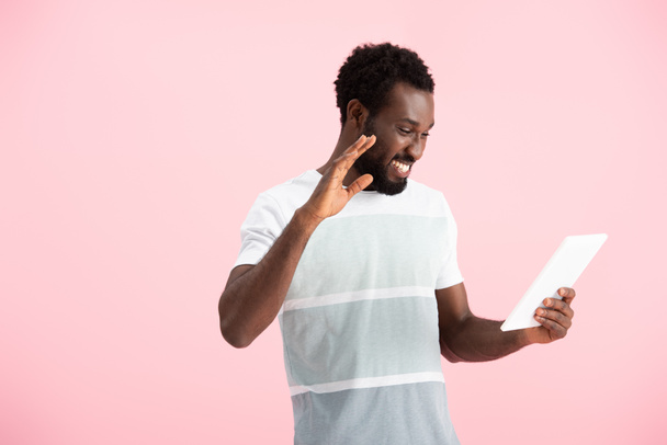 陽気なアフリカ系アメリカ人男性が手を振り、ピンクで隔離されたデジタルタブレット上でビデオチャットを持っている - 写真・画像