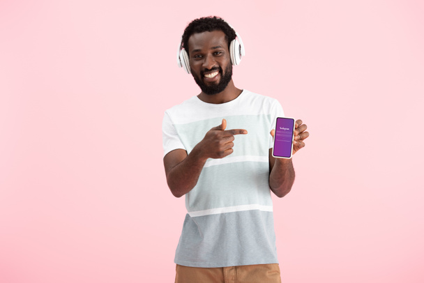 キエフ、ウクライナ - 2019年5月17日:アフリカ系アメリカ人男性がヘッドフォンで音楽を聴き、Instagramアプリでスマートフォンを指し示し、ピンクで隔離  - 写真・画像