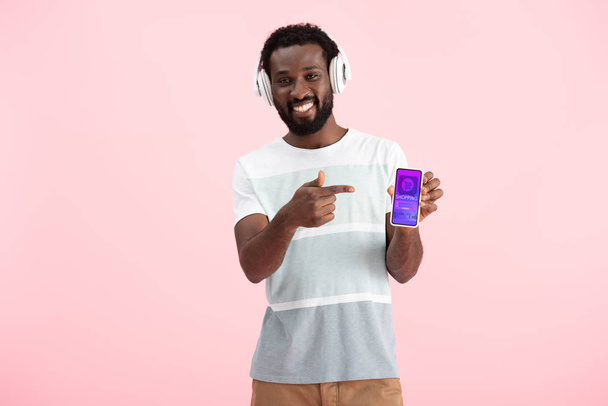 アフリカ系アメリカ人男性がヘッドフォンで音楽を聴き、ショッピングアプリでスマートフォンを指し示し、ピンクで隔離  - 写真・画像