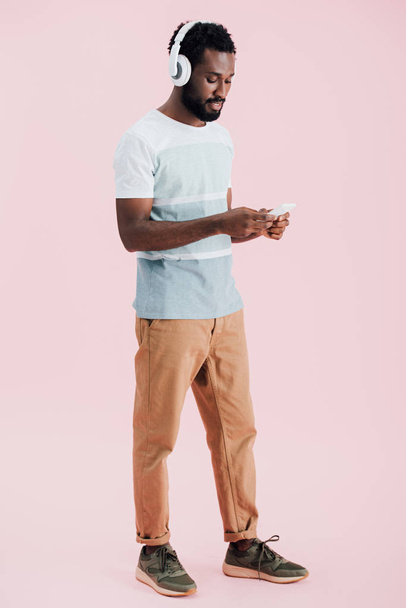 Afroameričan poslouchá hudbu se sluchátky a smartphone, izolovaný na růžovém - Fotografie, Obrázek