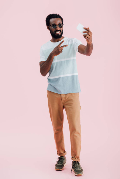Африканский американец в солнечных очках со знаком победы делает селфи на смартфоне, изолированный на розовом
  - Фото, изображение
