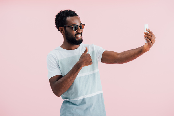 Afrikkalainen amerikkalainen mies aurinkolasit peukalo ylöspäin ottaen selfie älypuhelimeen, eristetty vaaleanpunainen
  - Valokuva, kuva