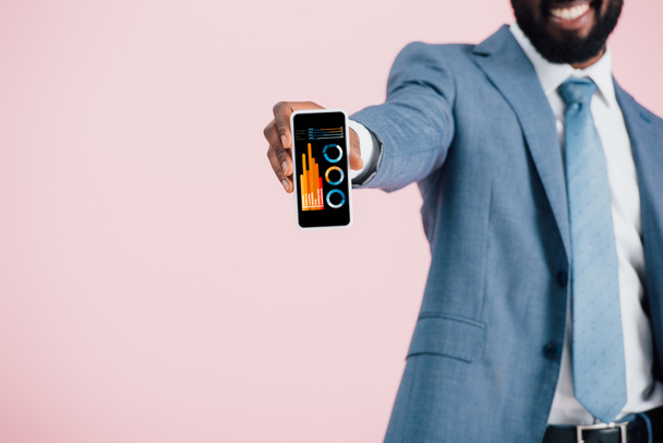 ピンクに隔離されたインフォグラフィックアプリでスマートフォンを示すスーツを着たアフリカ系アメリカ人ビジネスマンのトリミングビュー  - 写真・画像