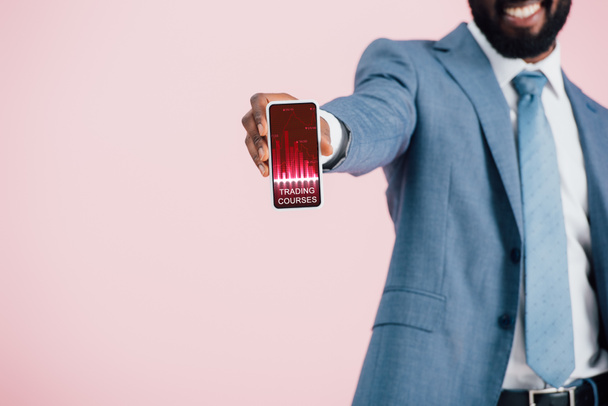貿易コースアプリでスマートフォンを示すスーツを着たアフリカ系アメリカ人ビジネスマンのトリミングビュー,ピンクで隔離  - 写真・画像