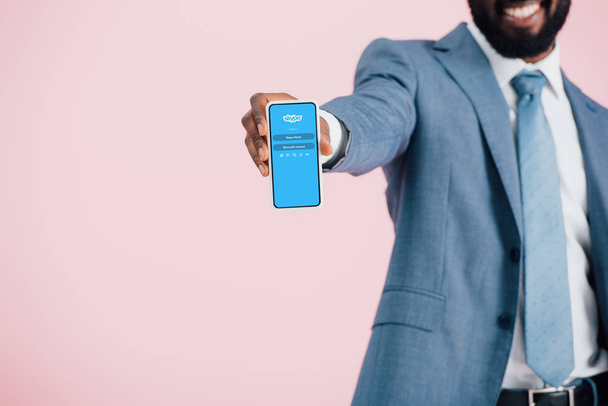 KYIV, UKRAINE - MAY 17, 2019: cropped view of african american businessman in suit showing smartphone with skype app, isolated on pink  - Fotografie, Obrázek