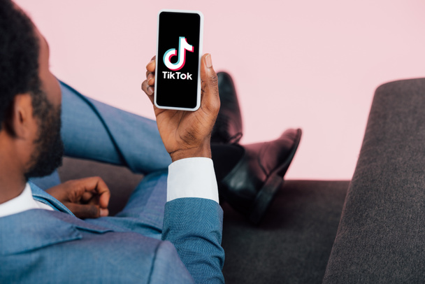 KYIV, UKRAINE - MAY 17, 2019: cropped view of african american businessman sitting on armchair and showing smartphone with Tik Tok app, isolated on pink - Foto, afbeelding