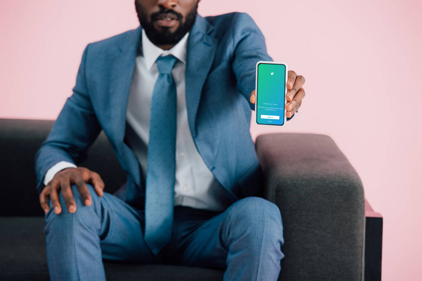 KYIV, UKRAINE - MAY 17, 2019: cropped view of african american businessman sitting on armchair and showing smartphone with twitter app, isolated on pink - Photo, Image