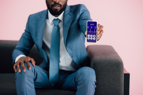обрезанный вид африканского американского бизнесмена, сидящего на кресле и показывающего смартфон с приложением для здравоохранения, изолированным на розовом
 - Фото, изображение