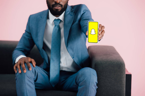КИЕВ, УКРАИНА - 17 мая 2019 года: обрезанный вид африканского американского бизнесмена, сидящего на кресле и показывающего смартфон с приложением Snapchat, изолированным на розовом
 - Фото, изображение