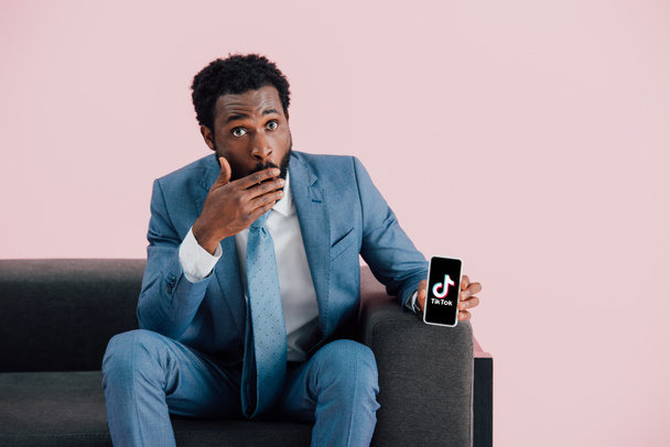 Κίεβο, Ουκρανία-17 Μαΐου 2019: σοκαρισμένος Αφρικανός Αμερικανός επιχειρηματίας σε κοστούμι κάθεται στην πολυθρόνα και δείχνοντας smartphone με την εφαρμογή tik tok, απομονώνονται σε ροζ - Φωτογραφία, εικόνα