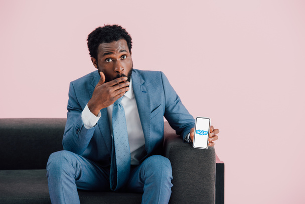 KYIV, UCRANIA - 17 de mayo de 2019: sorprendido hombre de negocios afroamericano en traje sentado en un sillón y mostrando el teléfono inteligente con aplicación skype, aislado en rosa
 - Foto, Imagen