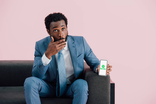 KYIV, UKRAINE - MAY 17, 2019: shocked african american businessman in suit sitting on armchair and showing smartphone with spotify app, isolated on pink - Foto, afbeelding