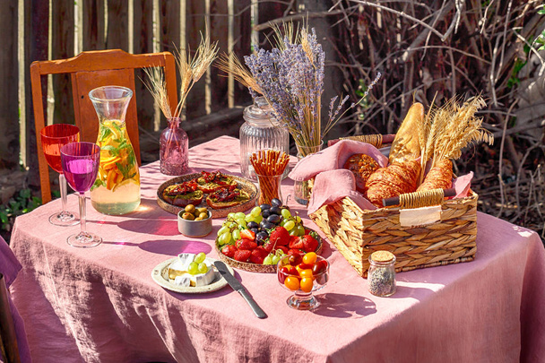 summer table with rose tablecloth with two colourful glasses and lemonade wattled basket with bread croissant and wheat outside sun rays bokeh lights - Photo, Image