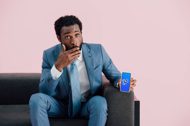 КИЕВ, Украина - 17 мая 2019 года: шокированный африканский бизнесмен в костюме сидит на кресле и показывает смартфон с приложением shazam, изолированным на розовом
 - Фото, изображение