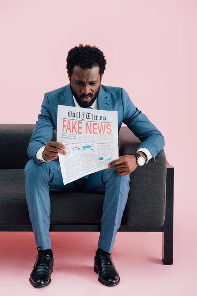 アフリカ系アメリカ人のビジネスマンがソファに座っている間にフェイクニュースで新聞を読んで動揺し、ピンクの上に孤立した  - 写真・画像