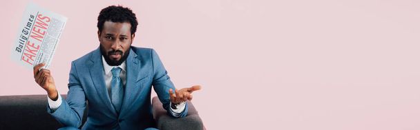 homme d'affaires afro-américain confus tenant journal avec de fausses nouvelles alors qu'il était assis sur le canapé, isolé sur rose
  - Photo, image