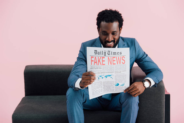 sorridente uomo d'affari afroamericano che legge giornali con notizie false mentre siede sul divano, isolato sul rosa
  - Foto, immagini