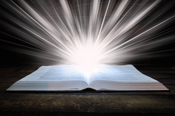 Raamatun suuri kirja on puupöydällä. Pimeässä. Kirjaan loistaa valo ylhäältä. Kirjasta tulee valoa.
 - Valokuva, kuva