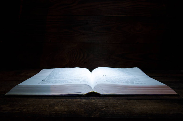 Το μεγάλο βιβλίο της Βίβλου βρίσκεται σε ένα ξύλινο τραπέζι. Στο σκοτάδι. Ένα φως λάμπει στο βιβλίο από πάνω. - Φωτογραφία, εικόνα