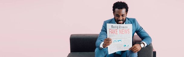 glimlachend African American zakenman lezing krant met nep nieuws terwijl zittend op de Bank, geïsoleerd op roze  - Foto, afbeelding