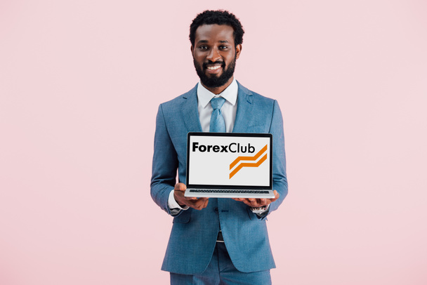 KYIV, UKRAINE - MAY 17, 2019: smiling african american businessman showing laptop with forex club website, isolated on pink - Fotografie, Obrázek