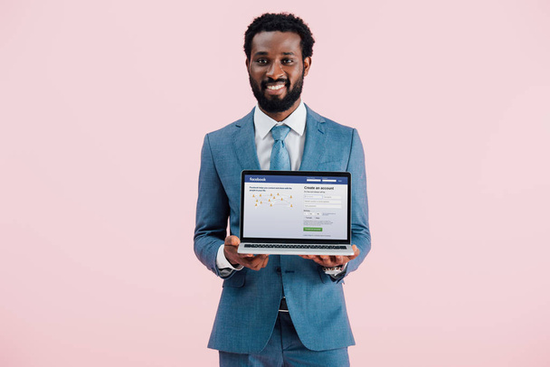 KYIV, UCRANIA - 17 DE MAYO DE 2019: sonriente hombre de negocios afroamericano mostrando portátil con el sitio web de facebook, aislado en rosa
 - Foto, imagen