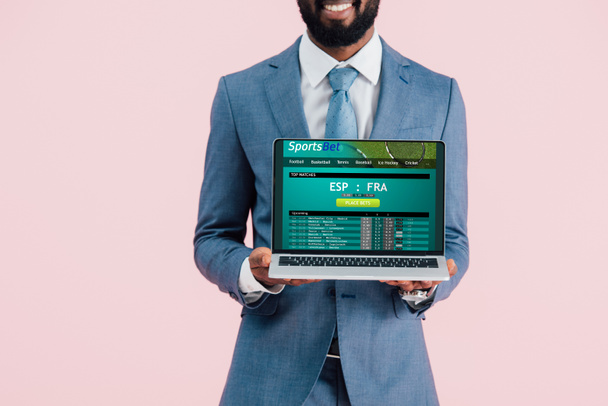青で隔離されたスポーツベットのウェブサイトでラップトップを示す笑顔のアフリカ系アメリカ人ビジネスマンのトリミングされたビュー - 写真・画像